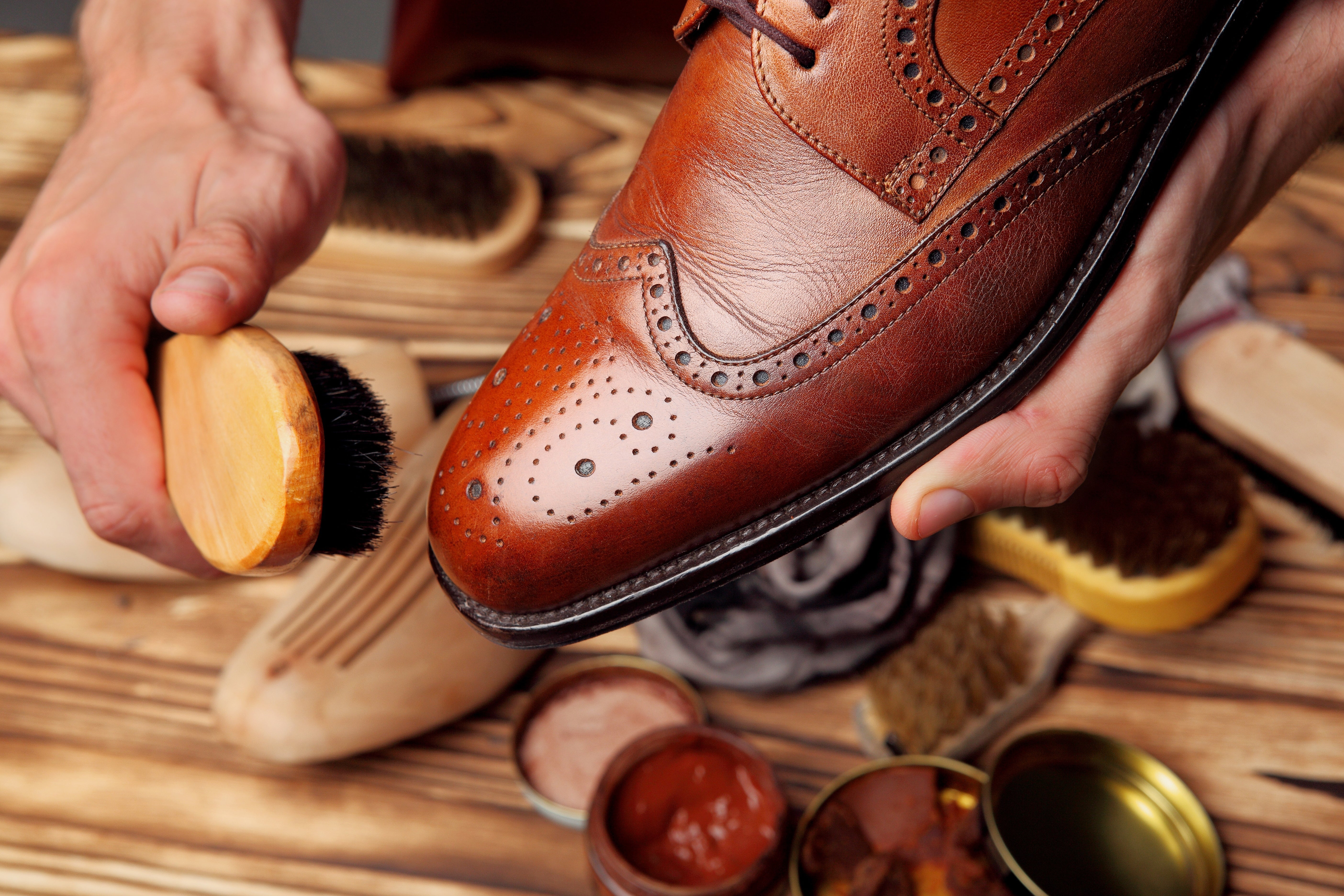 Hände reinigen einen braunen Lederschuh mit einer Schuhbürste, umgeben von Schuhcreme und Pflegezubehör auf einem Holztisch.